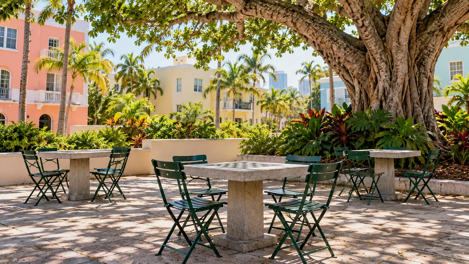 A small shaded park with empty domino tables and folding chairs under a large tree, pastel low buildings behind